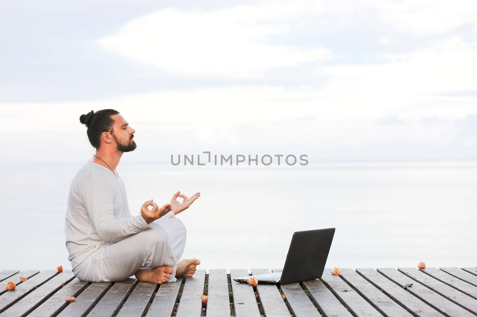 Caucasian man in white clothes meditating yoga with laptop on wooden pier