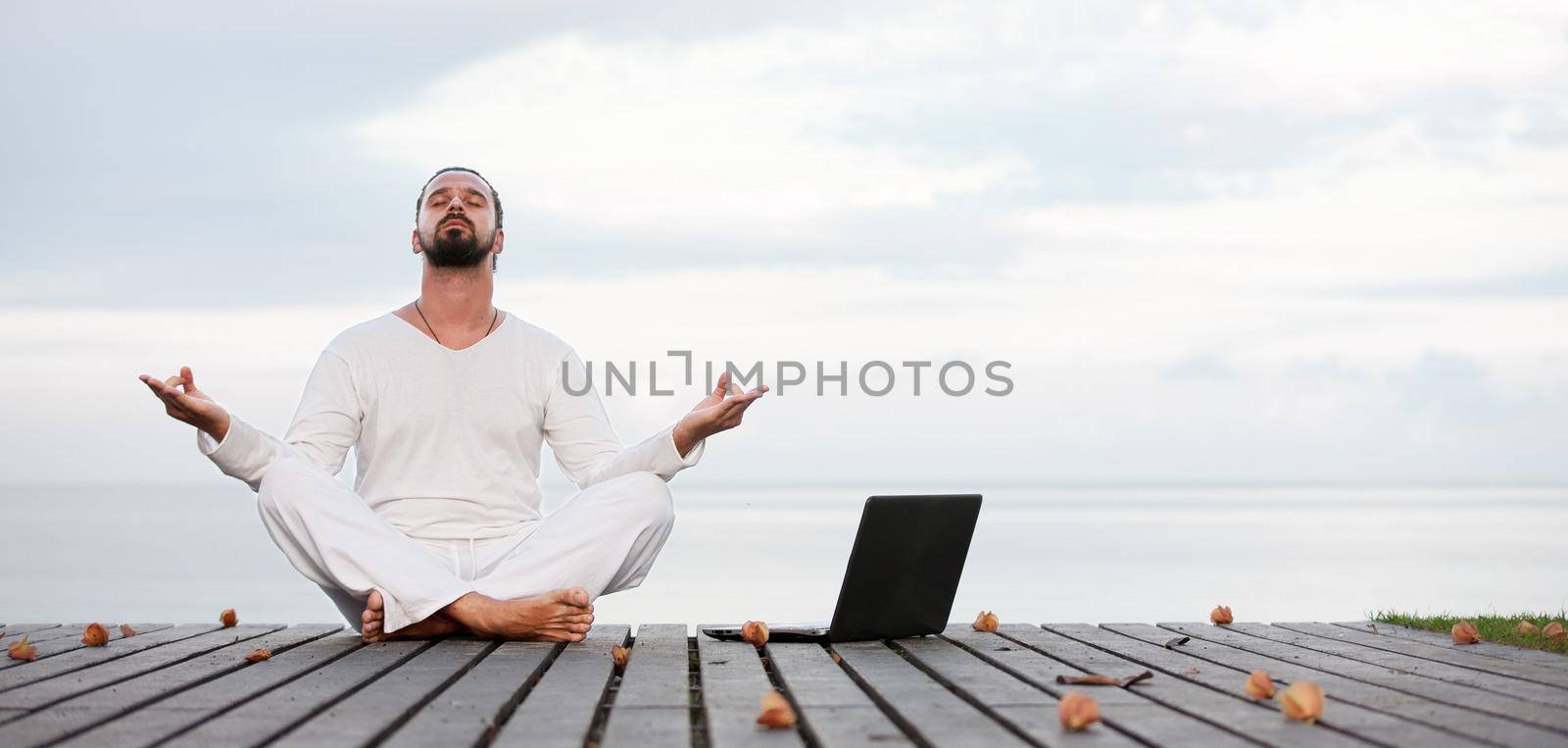 Caucasian man in white clothes meditating yoga with laptop on wooden pier