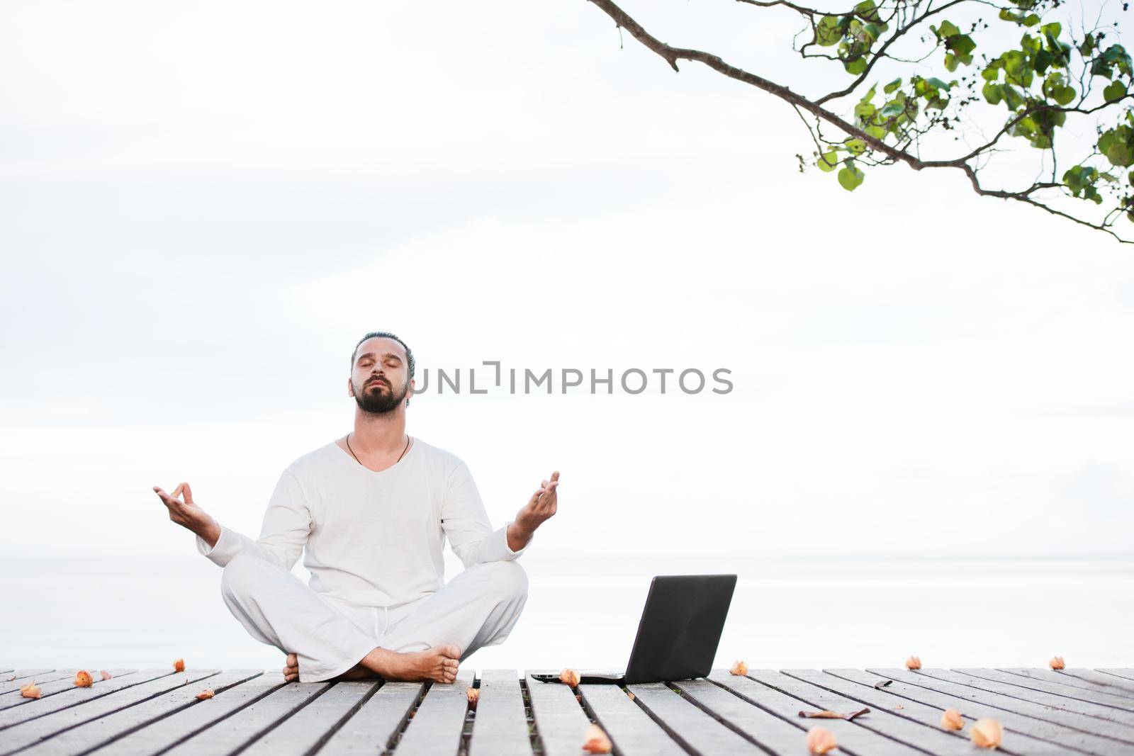 Caucasian man in white clothes meditating yoga with laptop on wooden pier