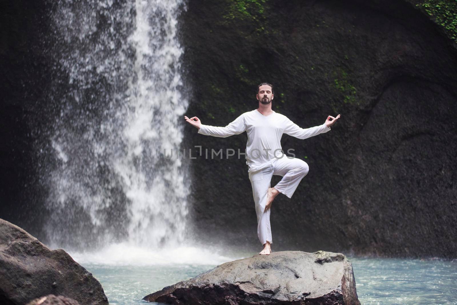 Man standing in meditation yoga on rock at waterfall by Jyliana