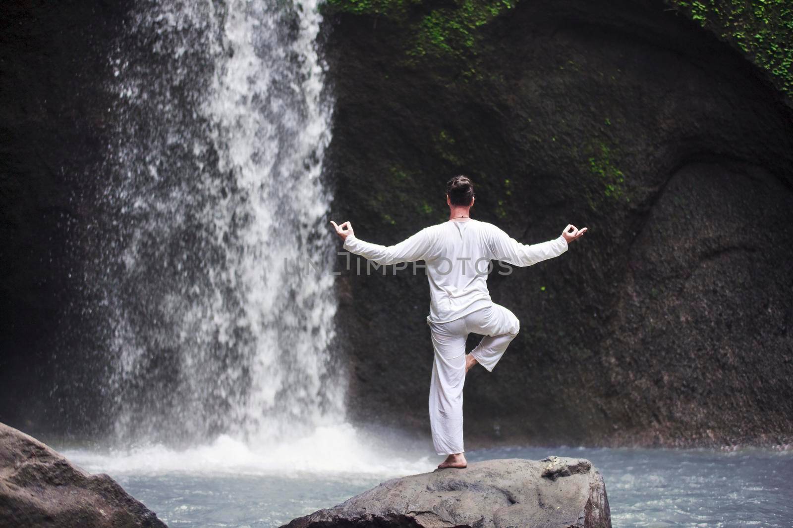 Man standing in meditation yoga on rock at waterfall by Jyliana