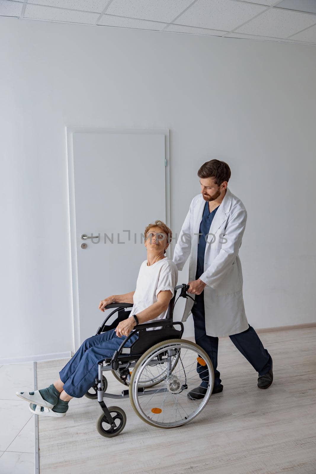 Professional male doctor carrying female patient on wheelchair in medicine clinic hall by Yaroslav_astakhov