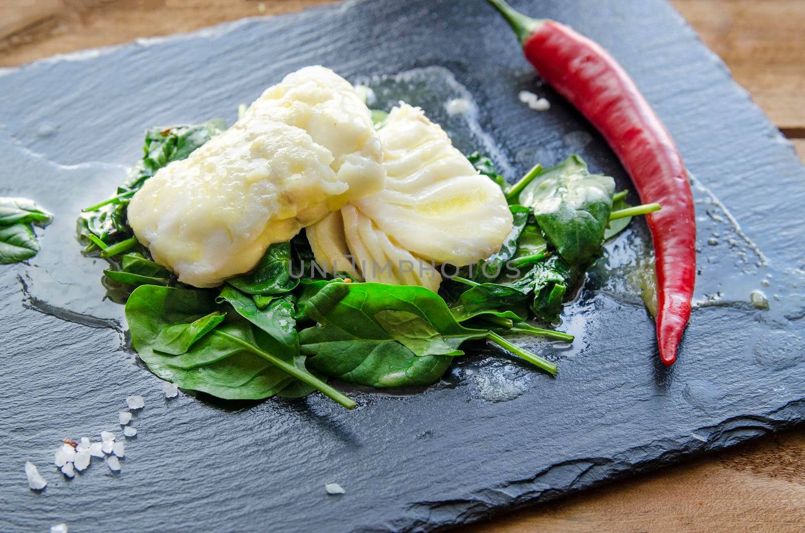 White fish fillet with herbs and chili on fried spinach. Gray background.