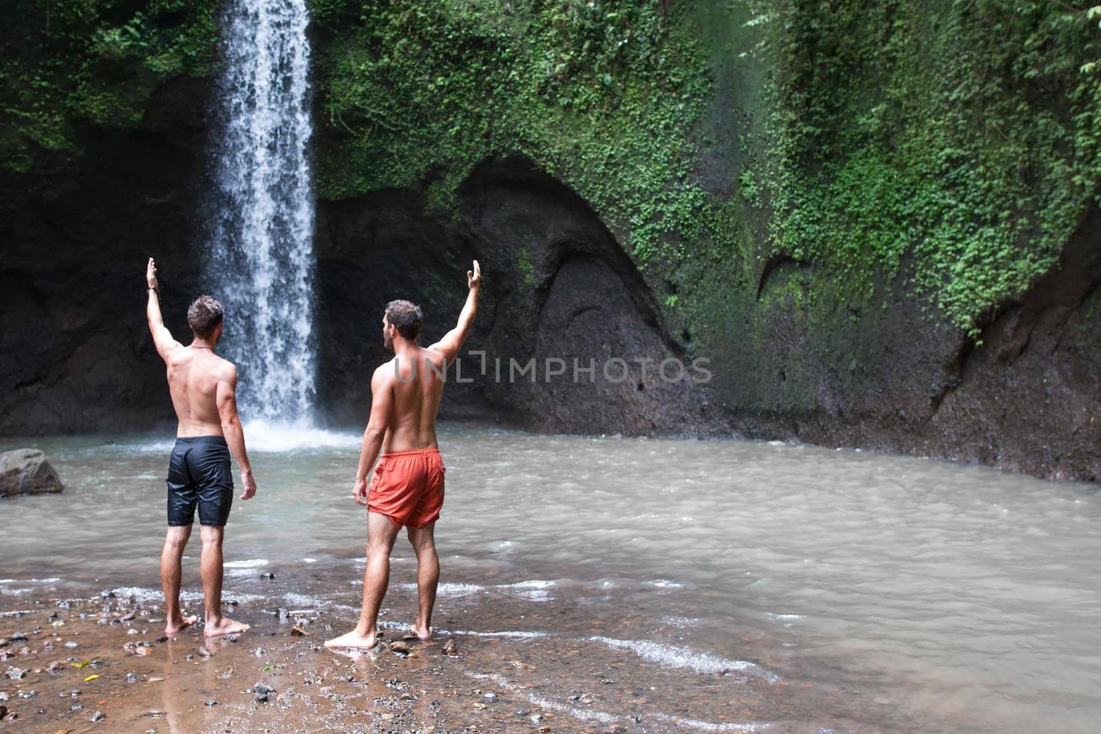 Two men with hands up near Beautiful Tibumana waterfall in bali by Jyliana