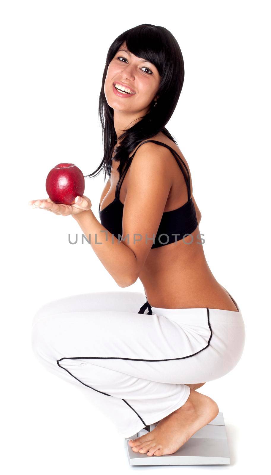 Happy young athlete woman standing on weighting scales and holding red apple isolated on white background