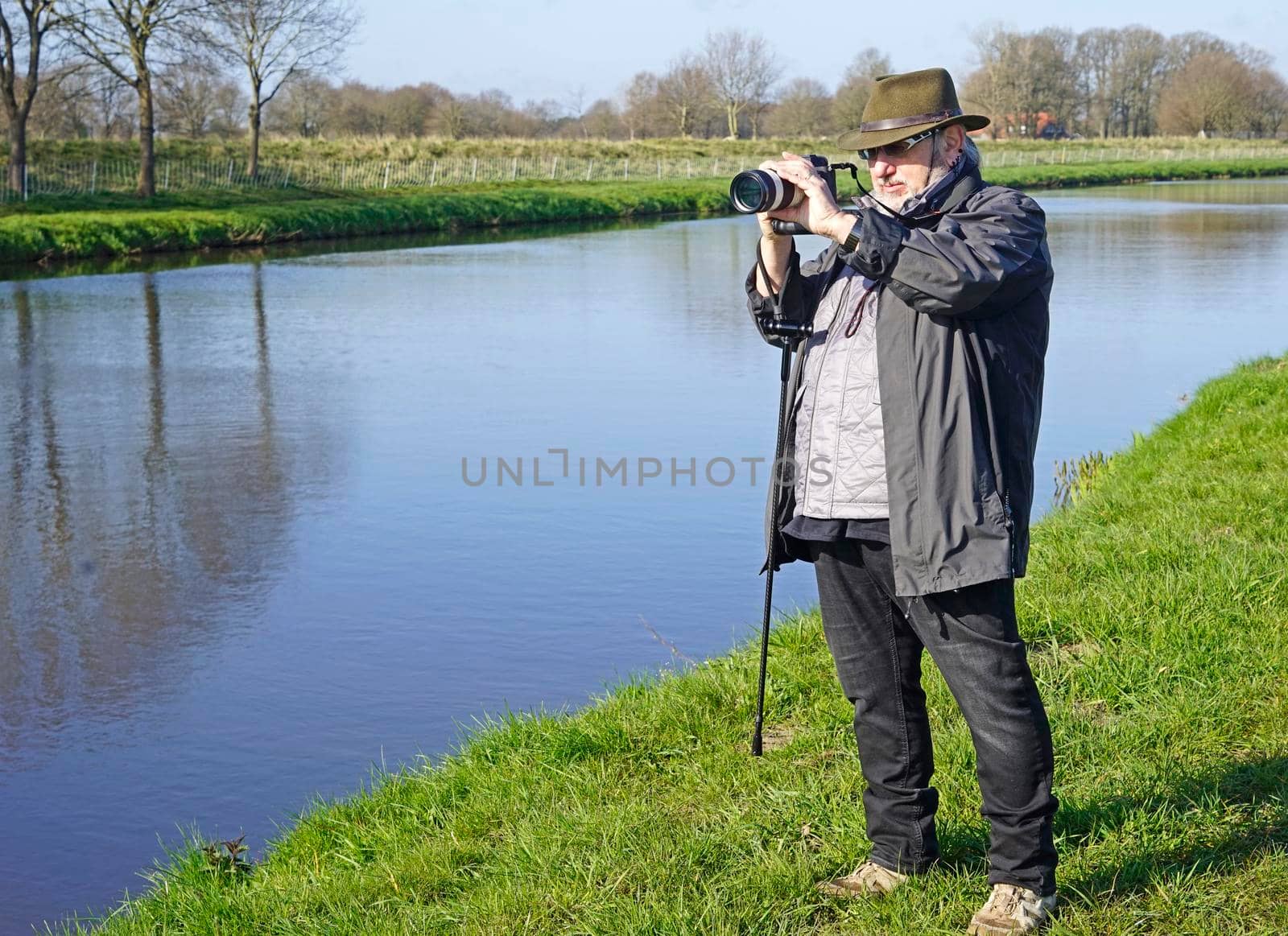 Senior photographer shooting a picture from a landscape near a river. The landscape is typical for  the Netherlands and this part of Germany: water, wide and flat