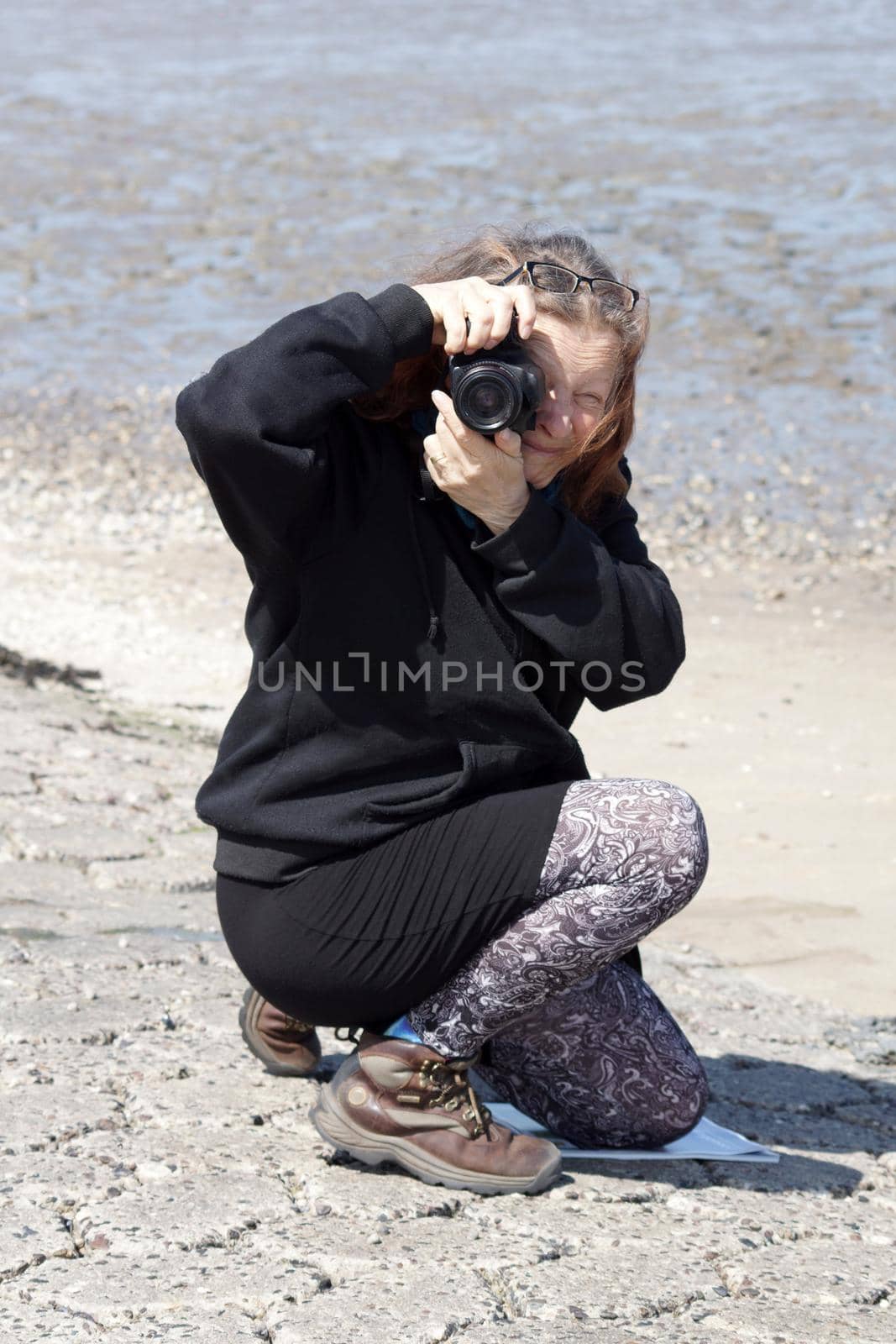 Elderly woman in black clothes photographs the photographer. She kneels on the sand and makes a vertical picture. Behind her, the silt of the Wadden Sea is visible
