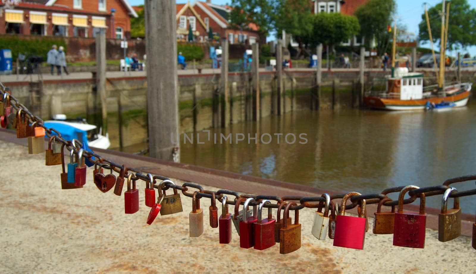 Love padlocks are attached to a rusted chain. In the background you can see the contours of an old fishing port out of focus. People walk on the quay.