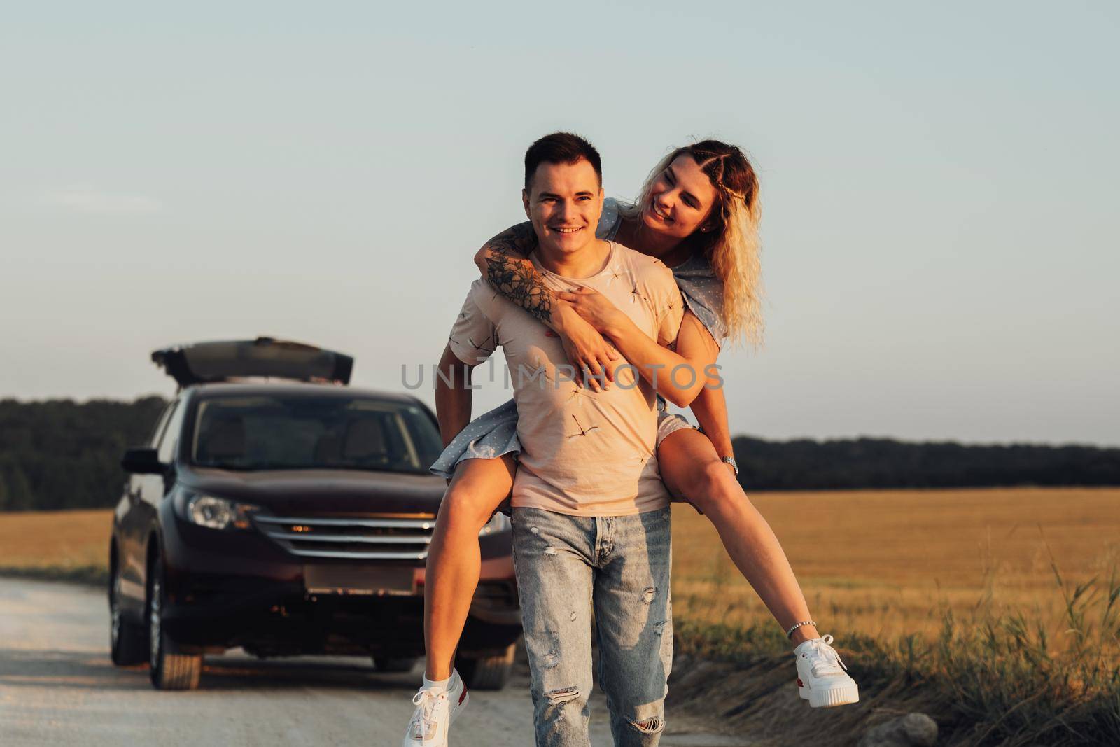 Caucasian Man Carries Girlfriend on His Back on Background of Their SUV Car, Young Couple Enjoying Their Road Trip at Sunset