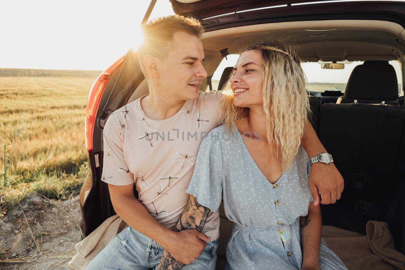 Young Woman and Man Sitting in Trunk of the Car, Happy Couple Enjoying Road Trip at Sunset by Romvy