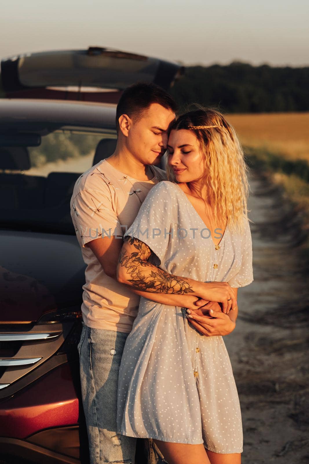Vertical Portrait of Cheerful Young Couple Hugging Near the Car, Tattooed Woman and Caucasian Man Enjoying Road Trip at Sunset by Romvy