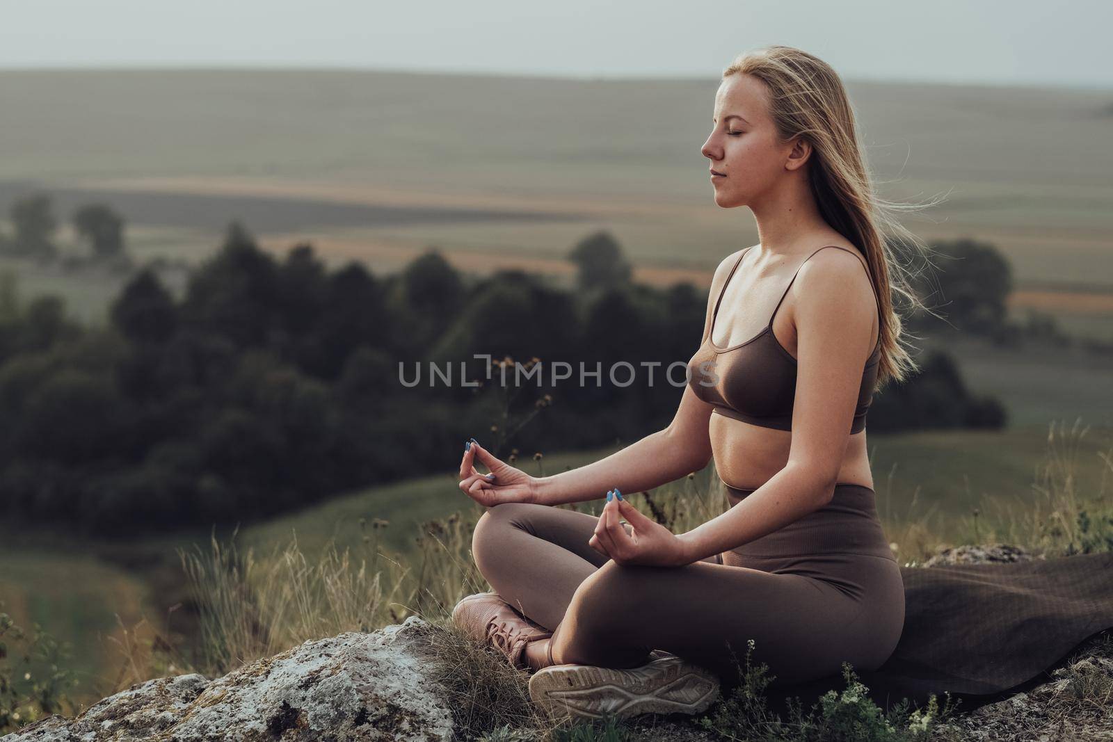 Sporty Girl Sitting in Lotus Pose and Doing Meditation Exercises on Top of a Hill, Young Woman Practice Yoga Outdoors with Panoramic Landscape at Sunset by Romvy