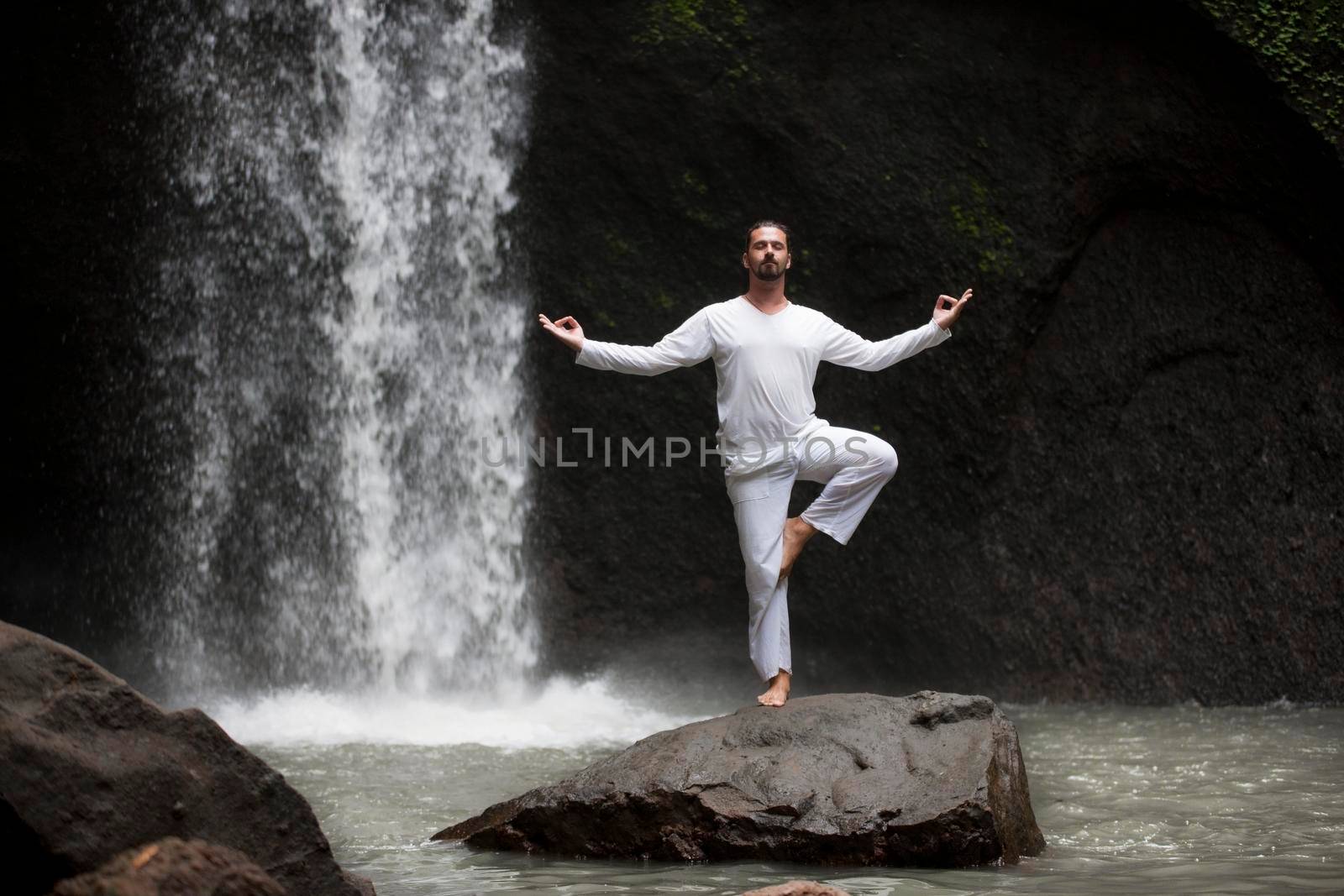 Man standing in meditation yoga on rock at waterfall by Jyliana