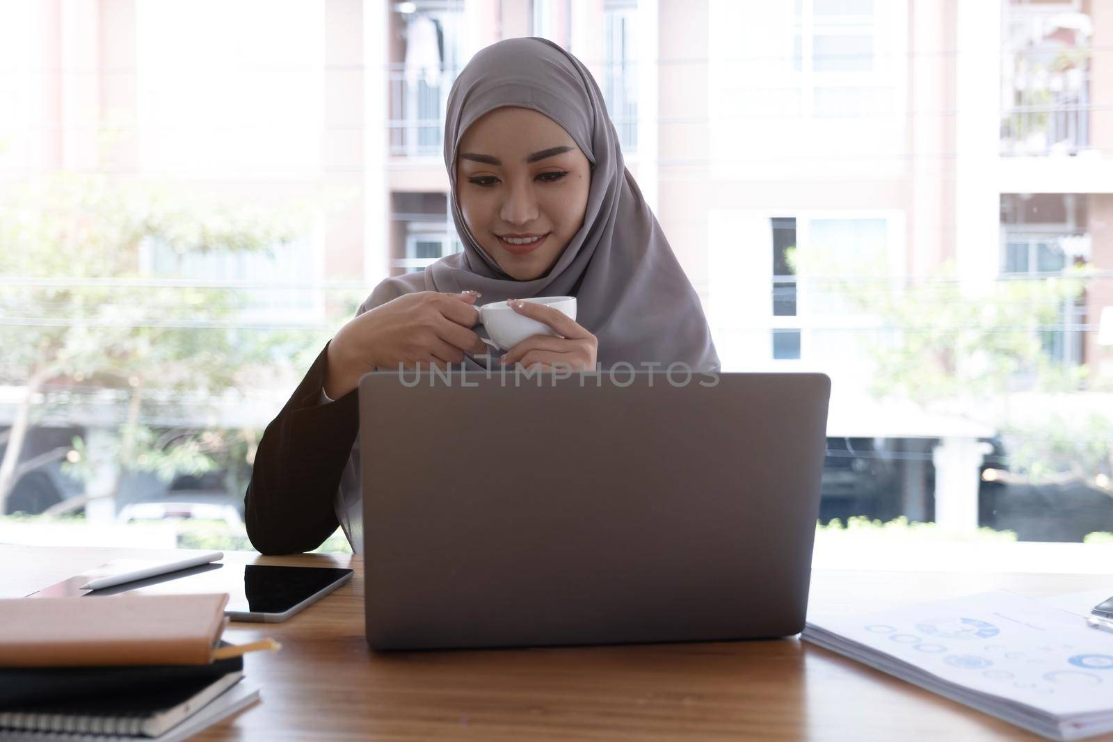 Asian Muslim hijab woman holding coffee cup looking at computer smiling happily by wichayada