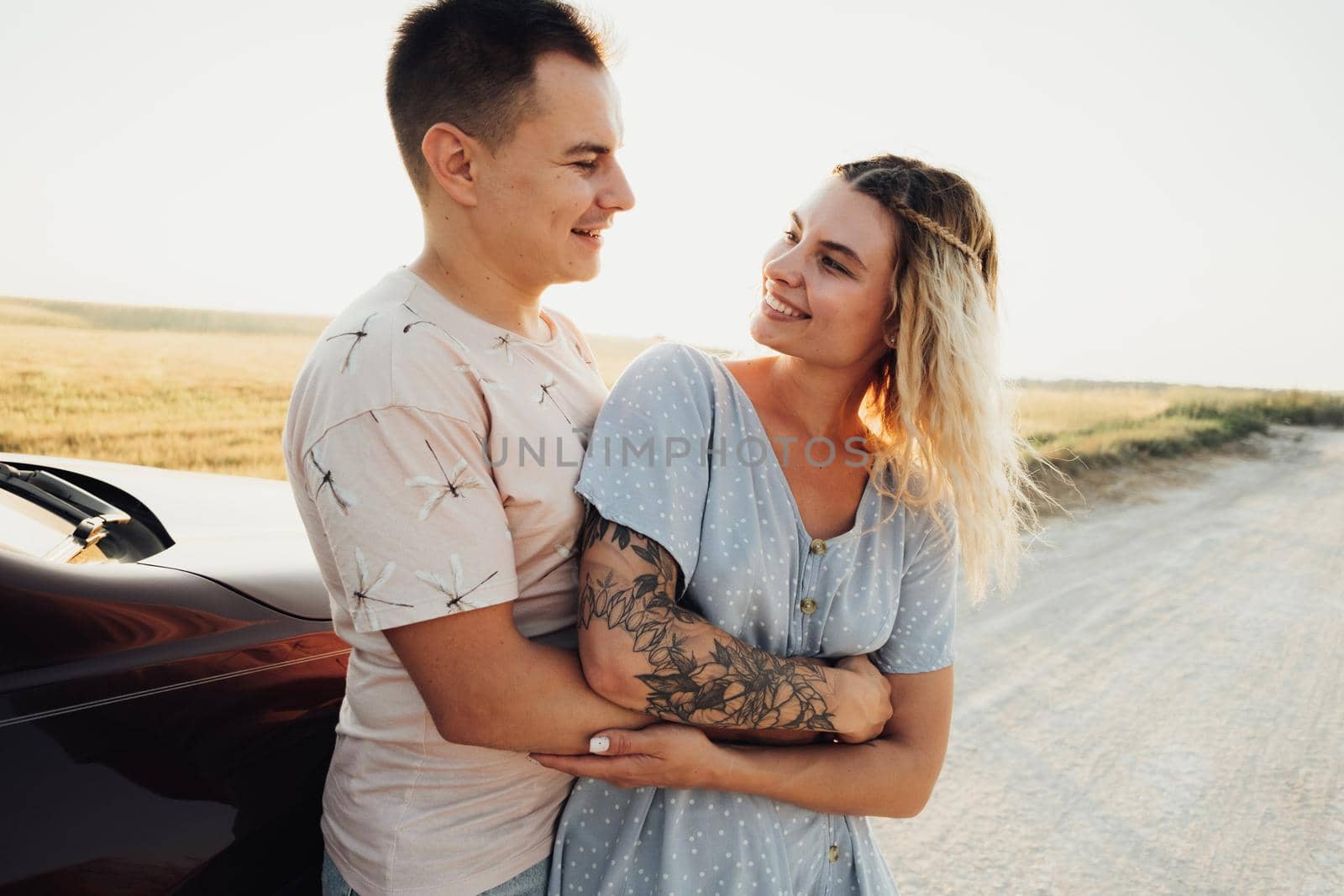 Cheerful Young Couple Hugging Near the Car, Tattooed Woman and Caucasian Man Enjoying Road Trip at Sunset by Romvy