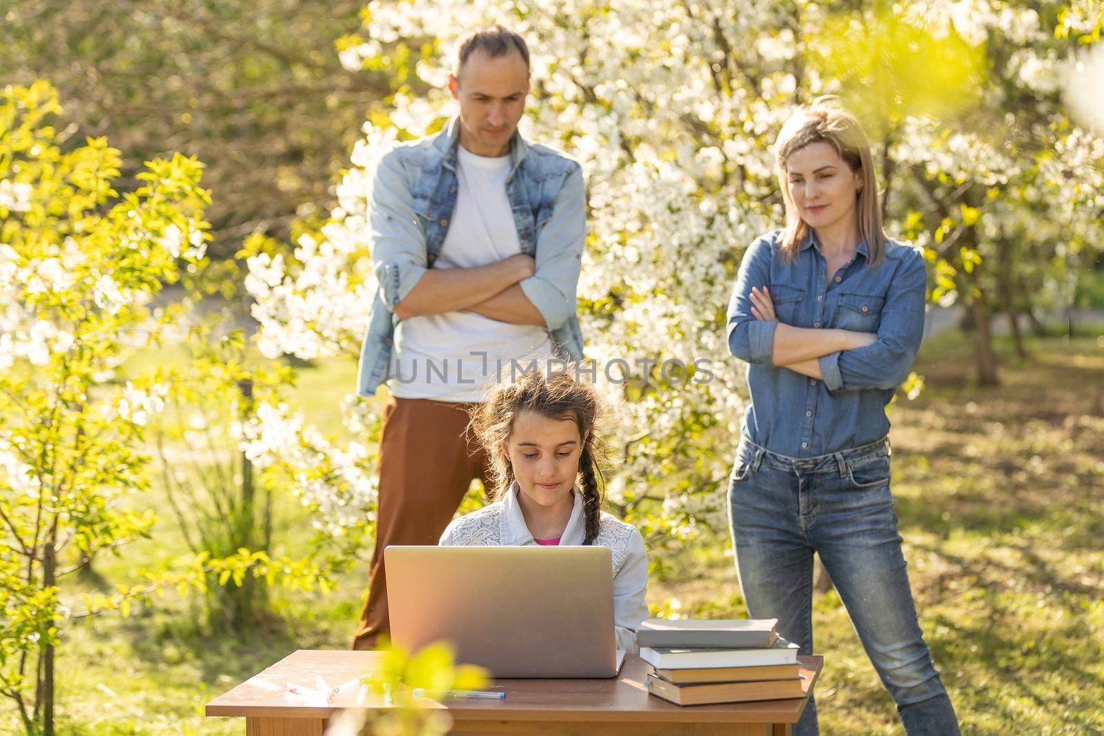 Happy family of four parents and cute little girl studying on laptop, enjoy using laptop watching cartoons, make internet video call or shopping online looking at computer screen sit together outdoor.