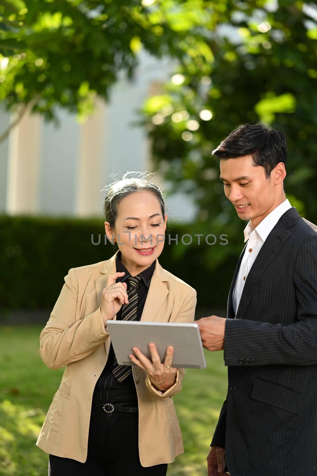 Mature female entrepreneur and her assistant discussing information on digital tablet while standing in front of modern office building by prathanchorruangsak