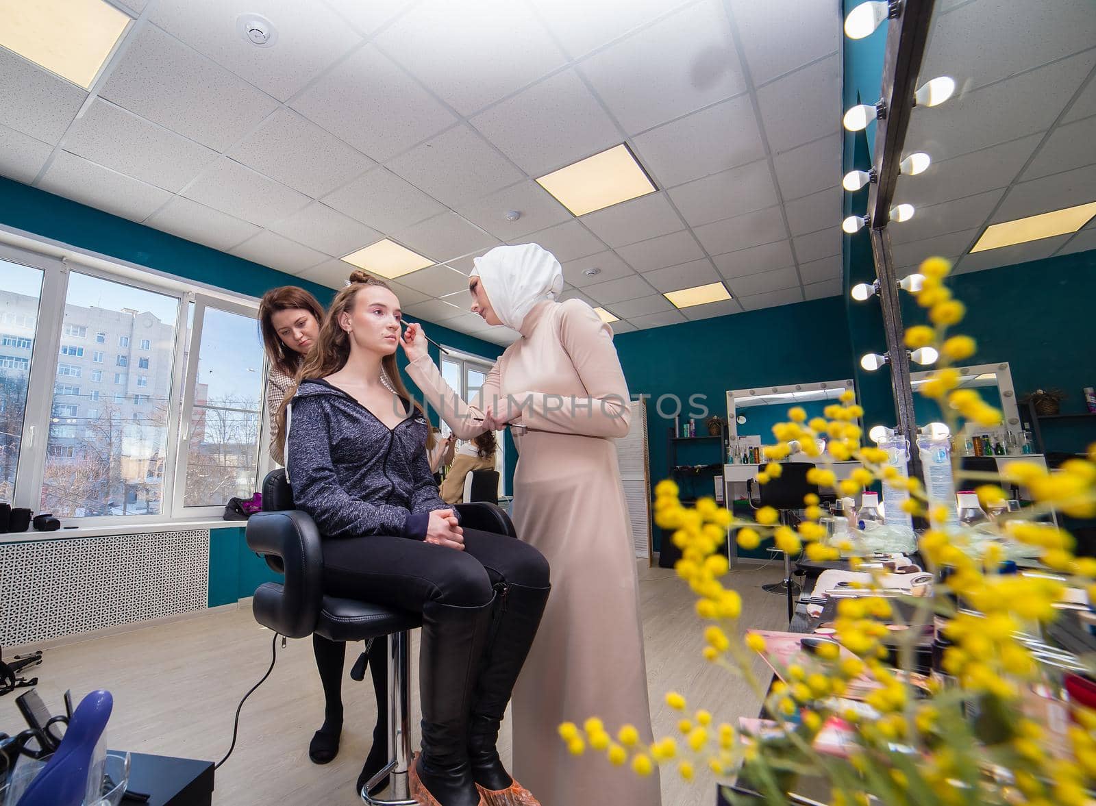 Two masters professionally style hair and make up a girl. Make-up artist and hairdresser create an image of a young woman. Business concept - beauty salon, facial skin and hair care.