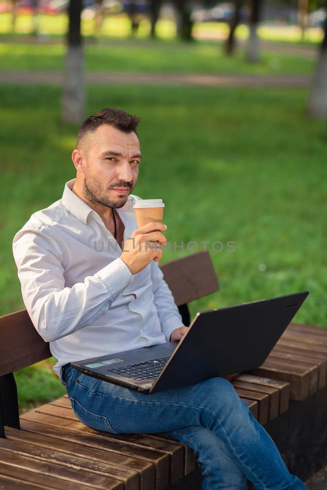 A man looking at the camera is working in the park with a laptop, drinking coffee. A young man on a background of green trees, a hot sunny summer day. Warm soft light, close-up.