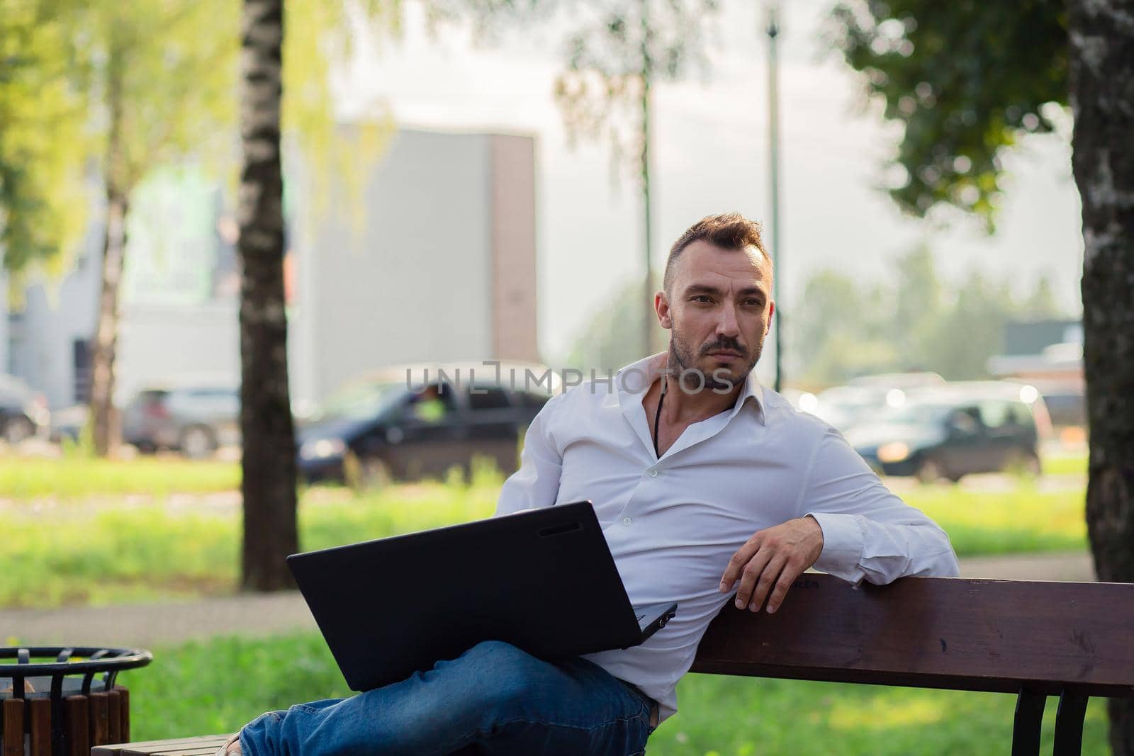 Thoughtful business man in a white shirt is working in the park with a laptop. Young man on the background of passing cars, hot sunny summer day. Warm soft light, close-up.