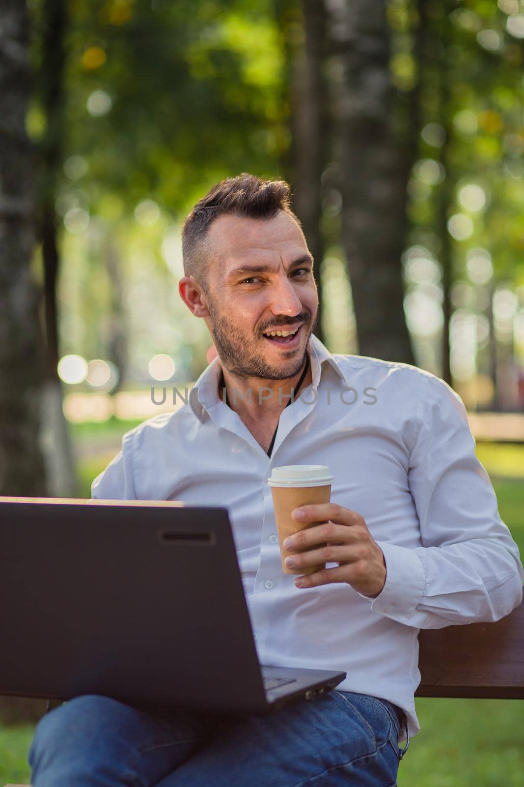 Laughing businessman is working in the park with a laptop, drinking coffee. A young man on a background of green trees, a hot sunny summer day. Warm soft light, close-up.