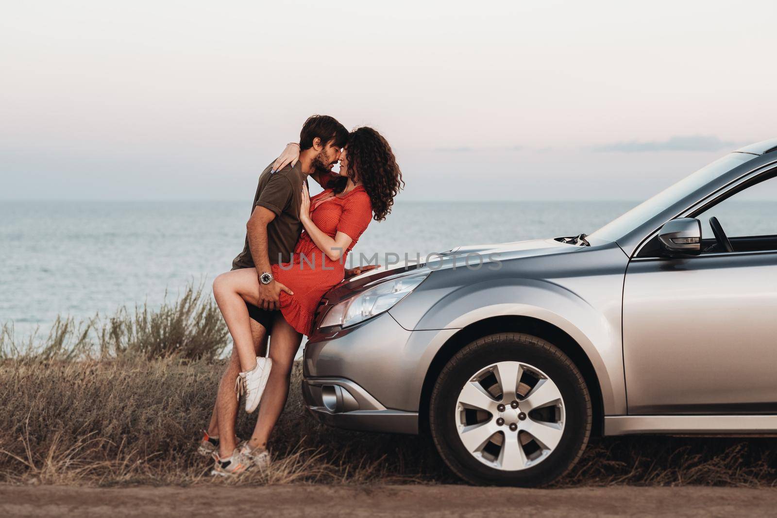 Young Lovers Man and Woman Hugging on the Hood of Their SUV Car on Background of Sea at Sunset