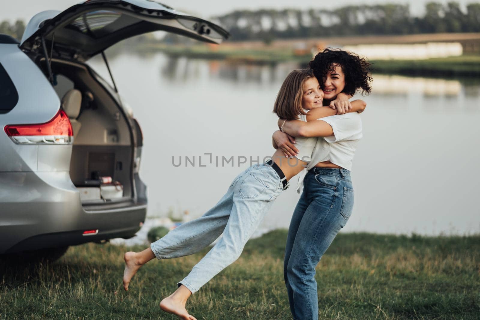 Happy Mother's Day, Woman Hugging with Cheerful Teenage Daughter Outdoors on the Background of Car and Lake