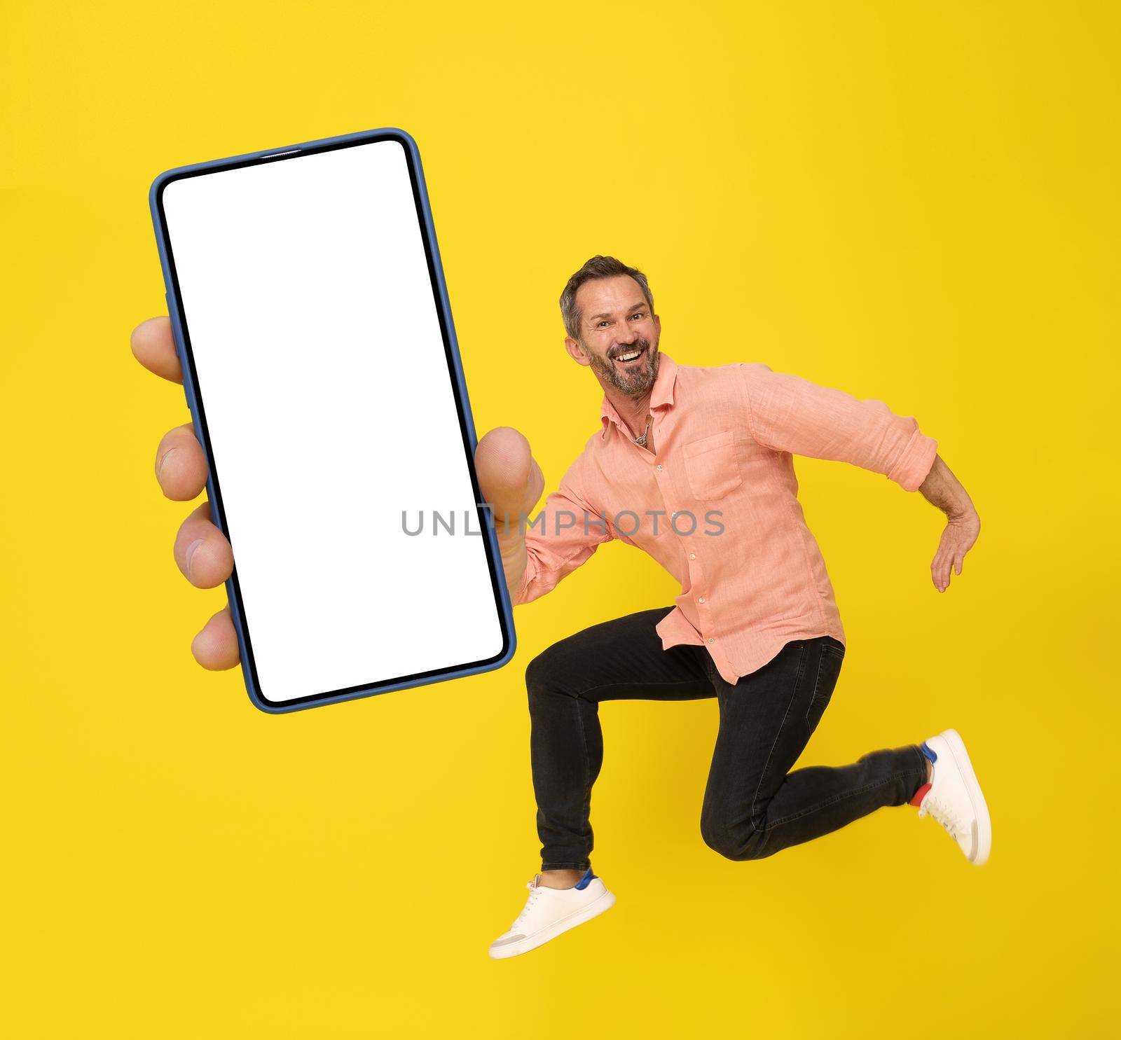 Mature grey haired man in high jump with smartphone in hand happy smiling on camera wearing peach shirt and black jeans isolated on yellow background. Mature fit man with smartphone.