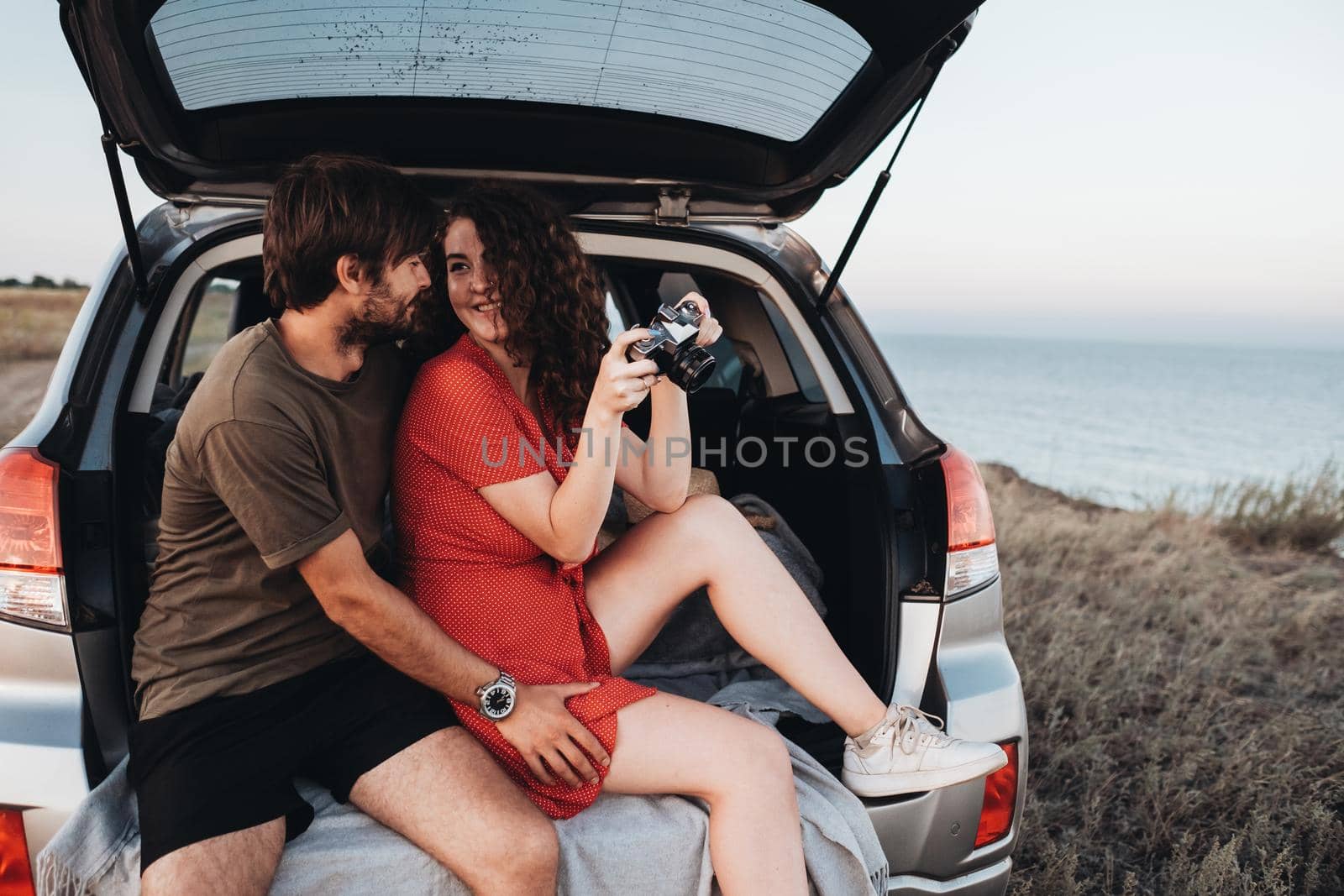 Man and Woman Sitting Inside Opened Trunk of SUV Car, Young Couple Enjoying Road Trip Along Sea at Sunset