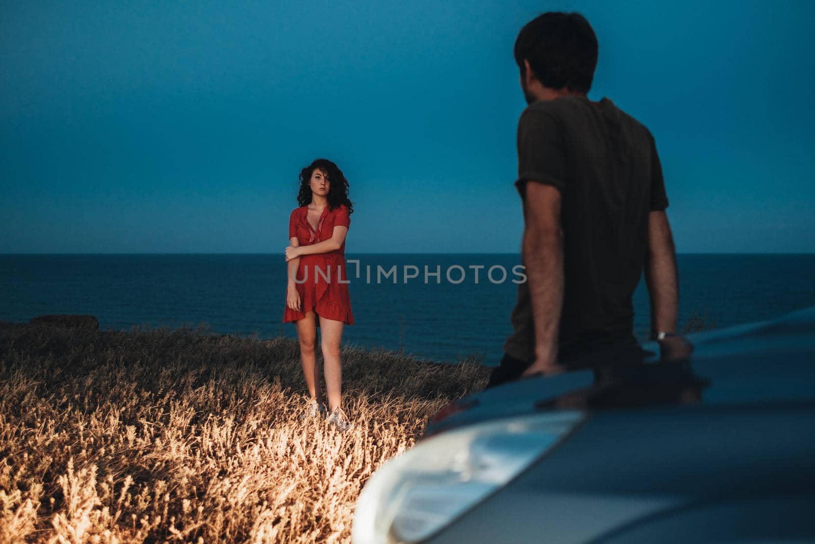 Man Sitting on the Hood of Car and Brunette Woman Dressed in Red Dress Standing in Front of Him Outdoors at Evening, Sea on Background
