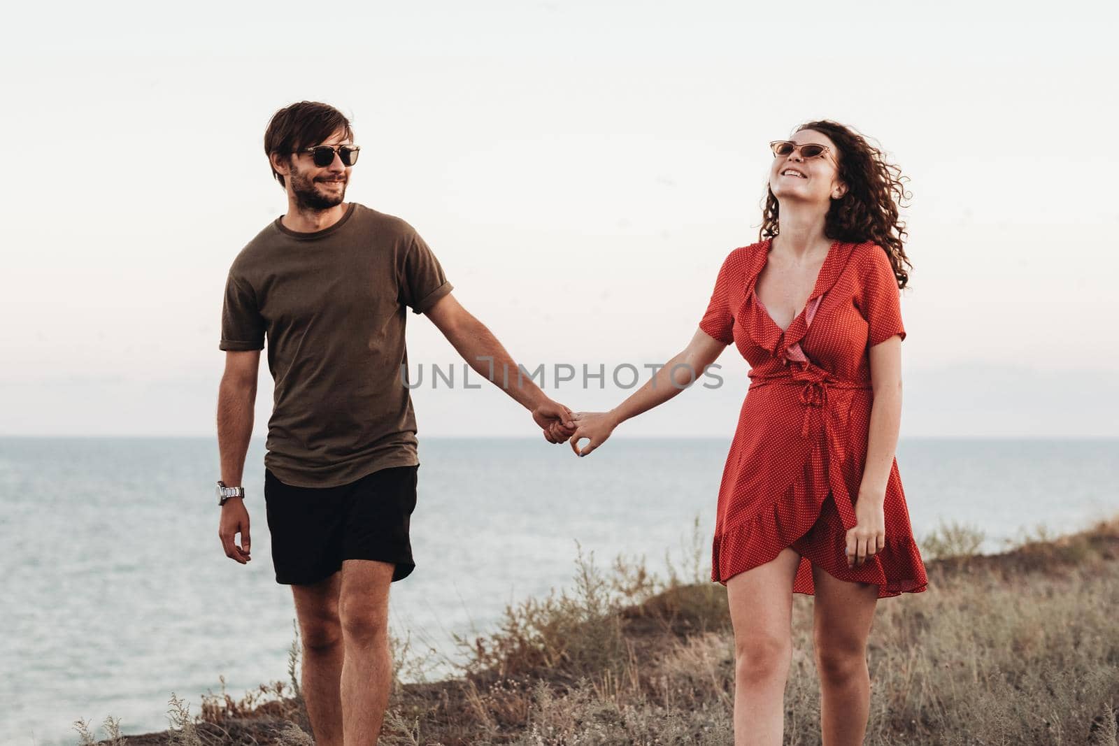 Cheerful Couple Walking Along Sea, Man and Curly Woman in Red Dress Holding by Hands Enjoying Their Vacation