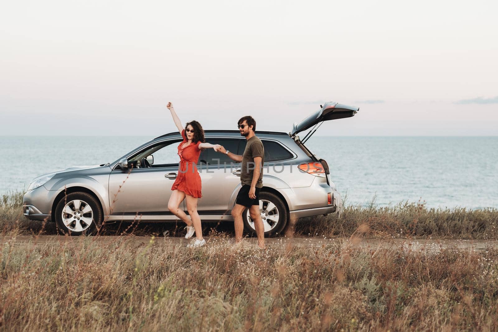 Youth Couple Having Fun on the Background of Sea, Man and Woman Enjoying Road Trip Near Their SUV Car at Sunset