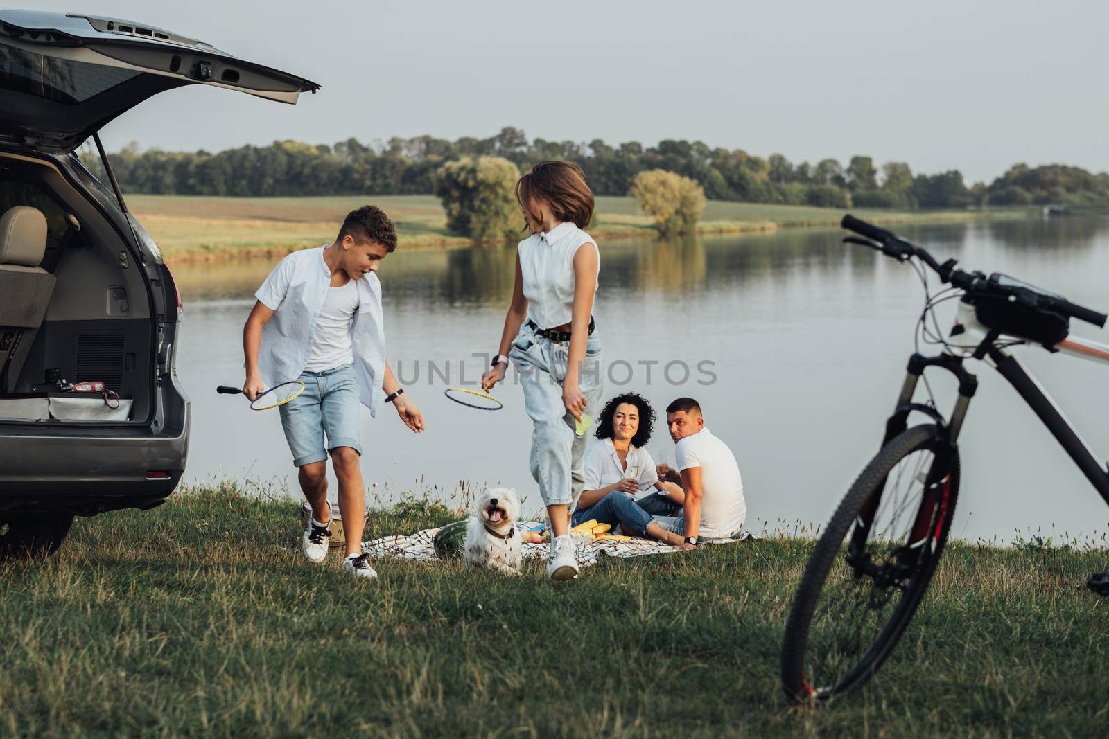 Two Teenage Children, Son and Daughter Playing with Pet Dog While Parents Having Picnic by Lake, Four Members Happy Family Enjoying Weekend Road Trip by Car