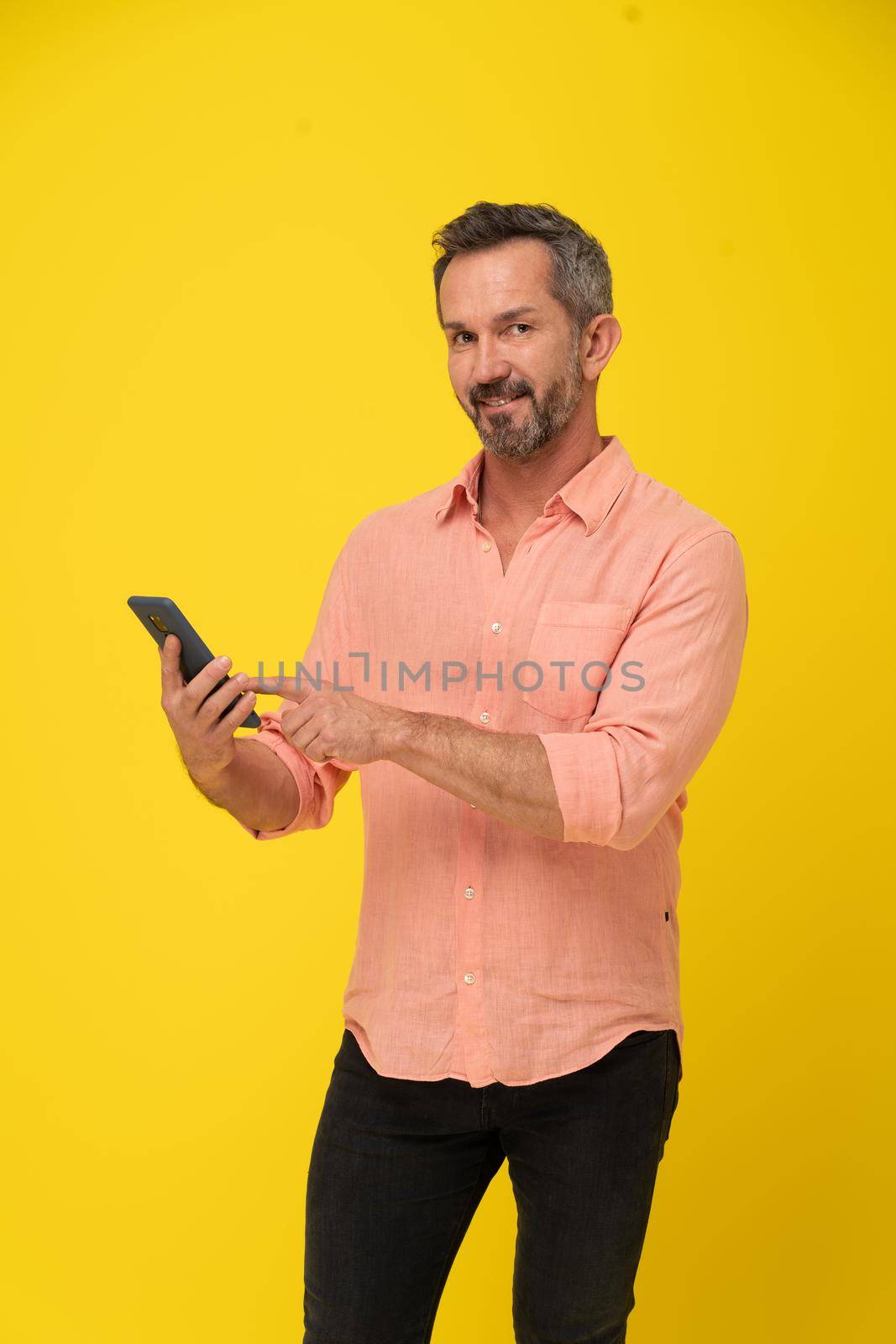 Grey haired man with smartphone in hand happy smiling on camera wearing peach shirt and black jeans isolated on yellow background. Mature fit man with smartphone.