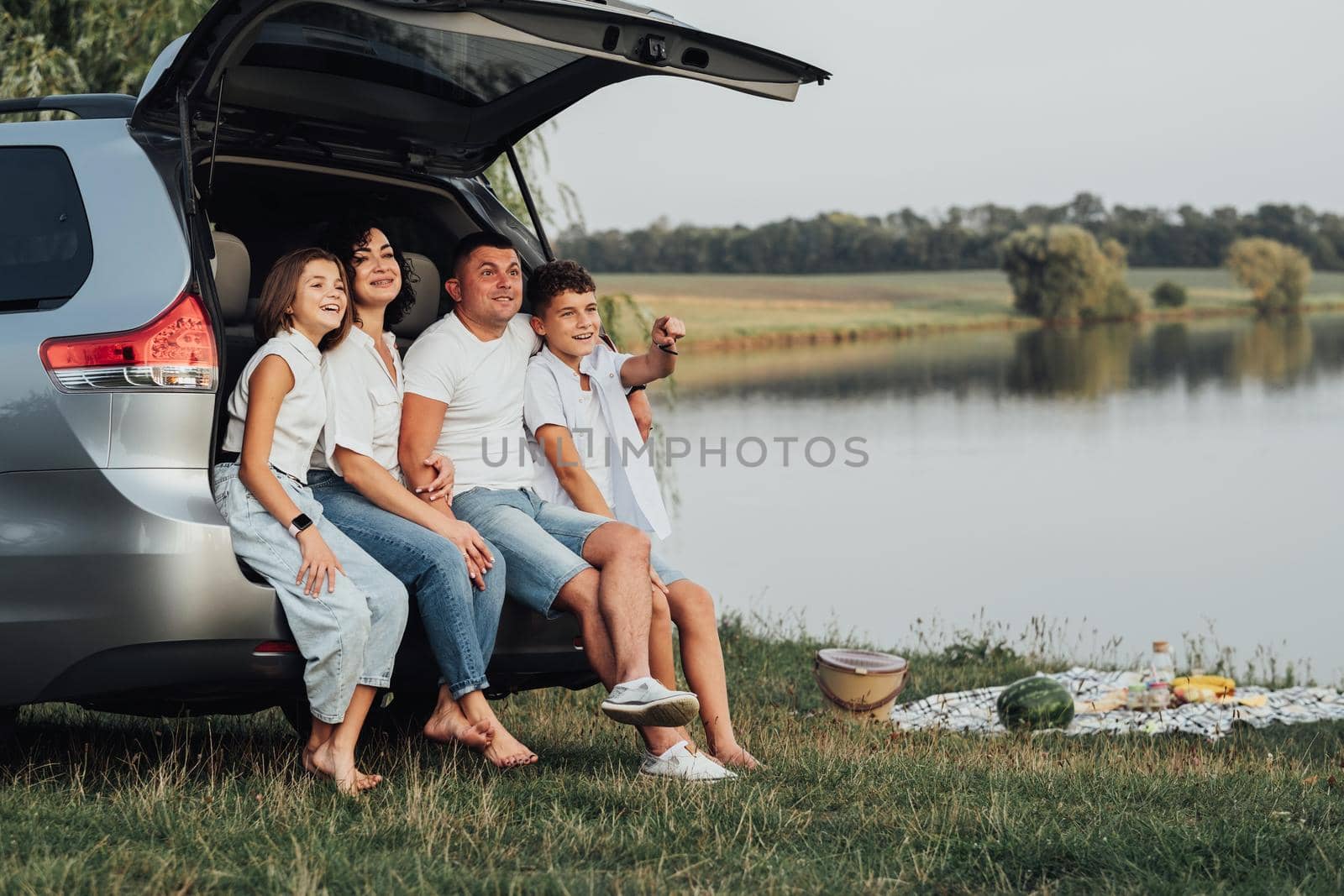 Happy Four Caucasian Members Family Sitting in Trunk of Minivan Car, Mother and Father with Two Teenage Children, Son and Daughter Having Weekend Picnic Outdoors by the Lake by Romvy
