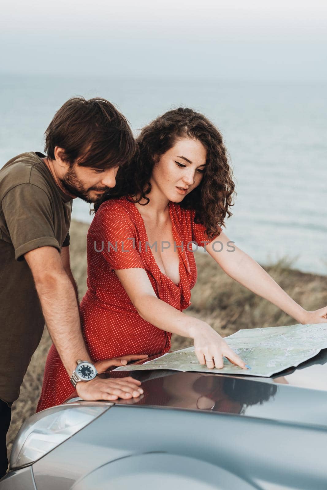 Happy Young Couple Man and Woman Checking the Map on Hood of Their Car to Plan Their Next Road Trip Along Seaside by Romvy