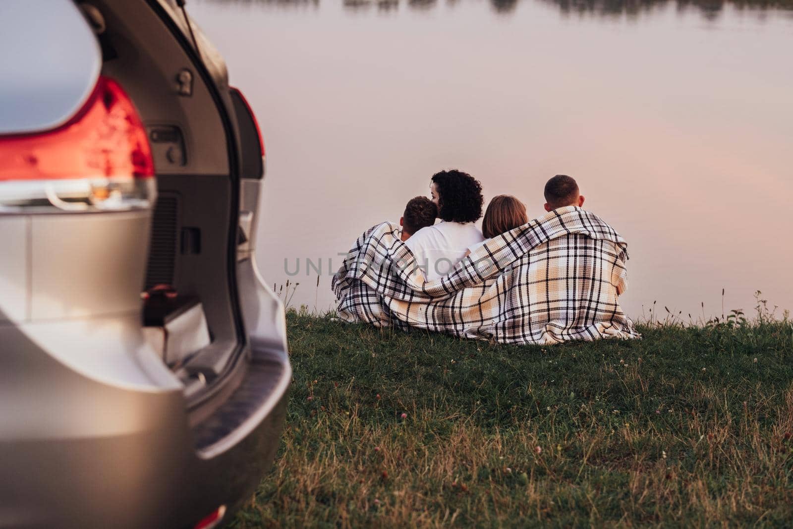 Four Members Family Sitting on the Grass by the Lake at Sunset, Mother and Father with Two Children Enjoying Weekend Road Trip by Romvy