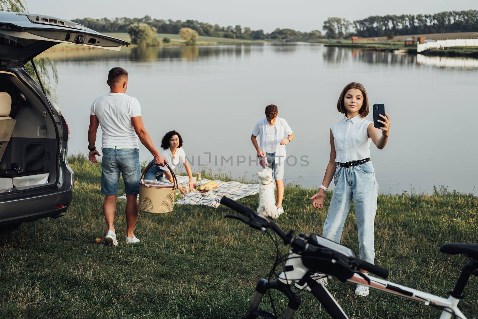 Happy Four Members Family Enjoying Weekend Road Trip by Minivan Car, Teenage Daughter Making Selfie While Her Brother Playing with Pet Dog by the Lake by Romvy