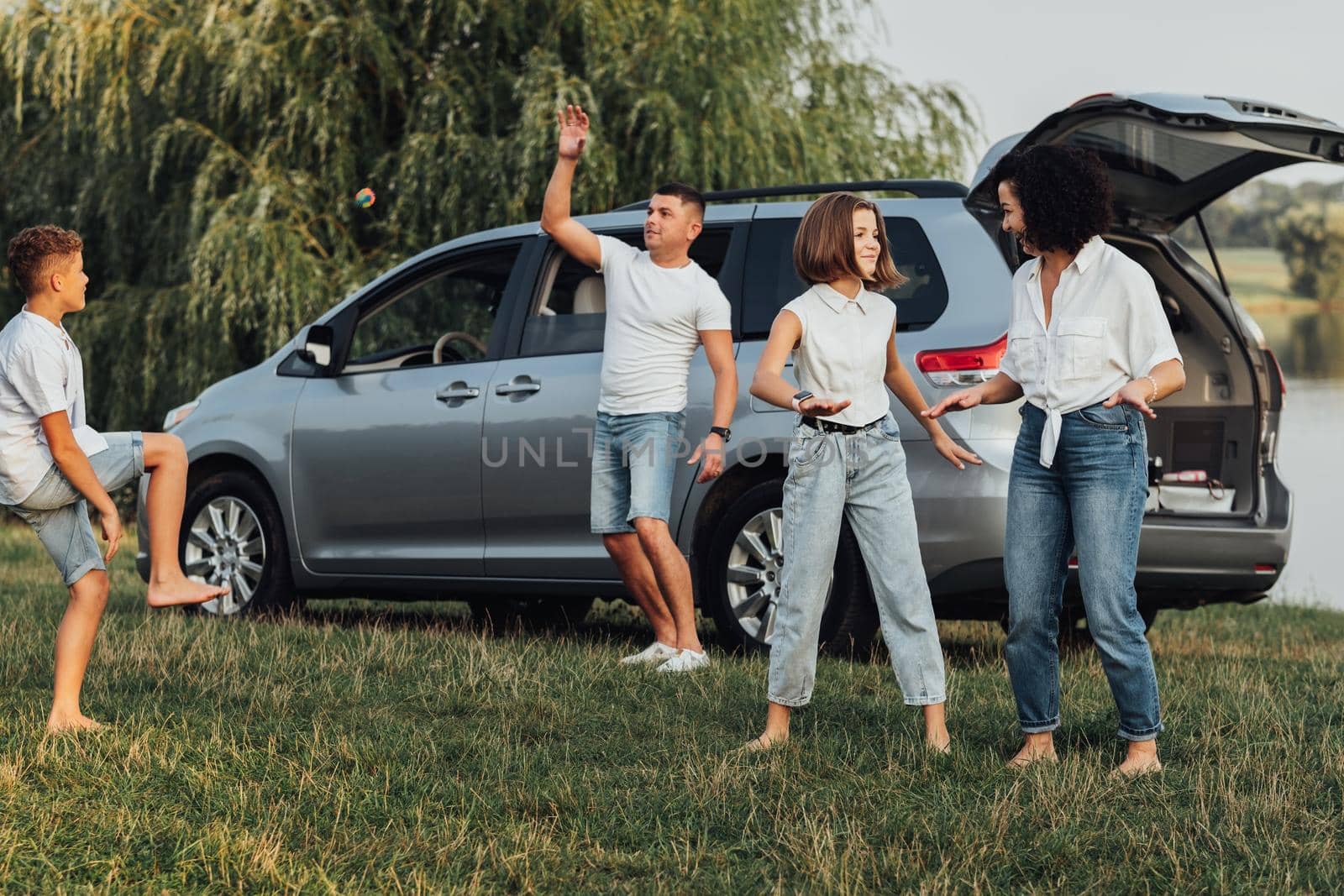 Happy Four Members Family Having Fun Time Outdoors, Mother Dancing with Teenage Daughter While Father Playing with Son, Minivan Car on the Background by Romvy