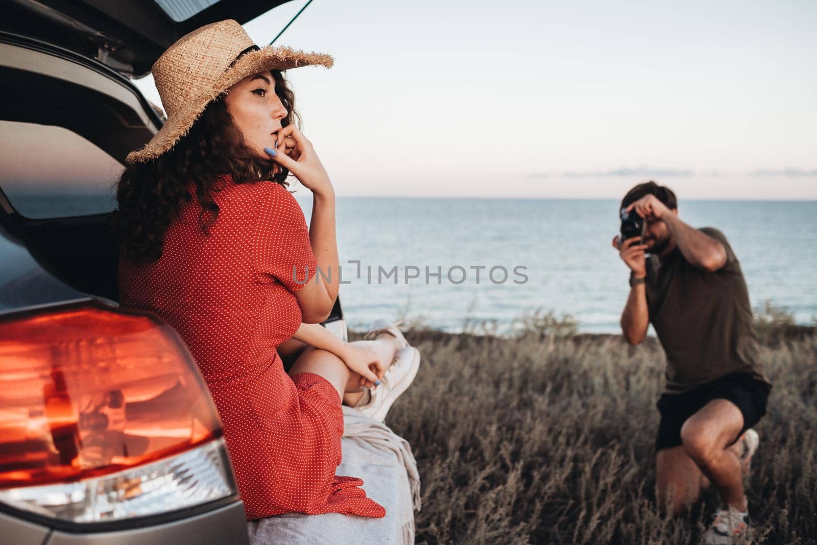 Man Photographing Woman in Red Dress and Hat That Sitting Inside Opened Trunk of Car on the Background of Sea