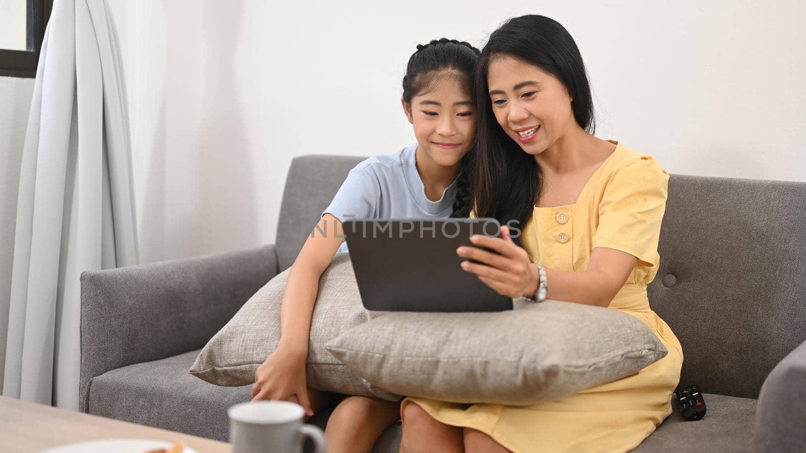 Smiling asian mom and daughter having fun browsing internet on digital tablet, enjoying spending free time together.