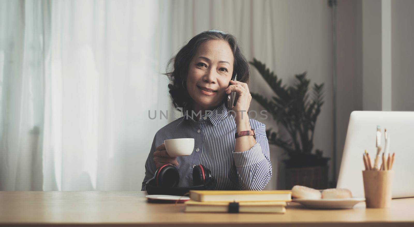 Happy pretty mature woman speaking on mobile phone portrait. Senior grey haired lady making telephone call, sitting on couch at home, enjoying talk, communication, looking at camera, smiling by wichayada