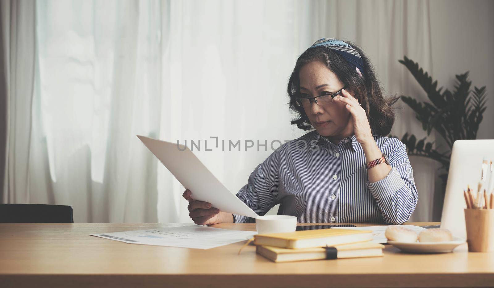 Seriously senior woman tworking at home, looking at laptop and waving paper correspondence, sitting at home, free space. by wichayada