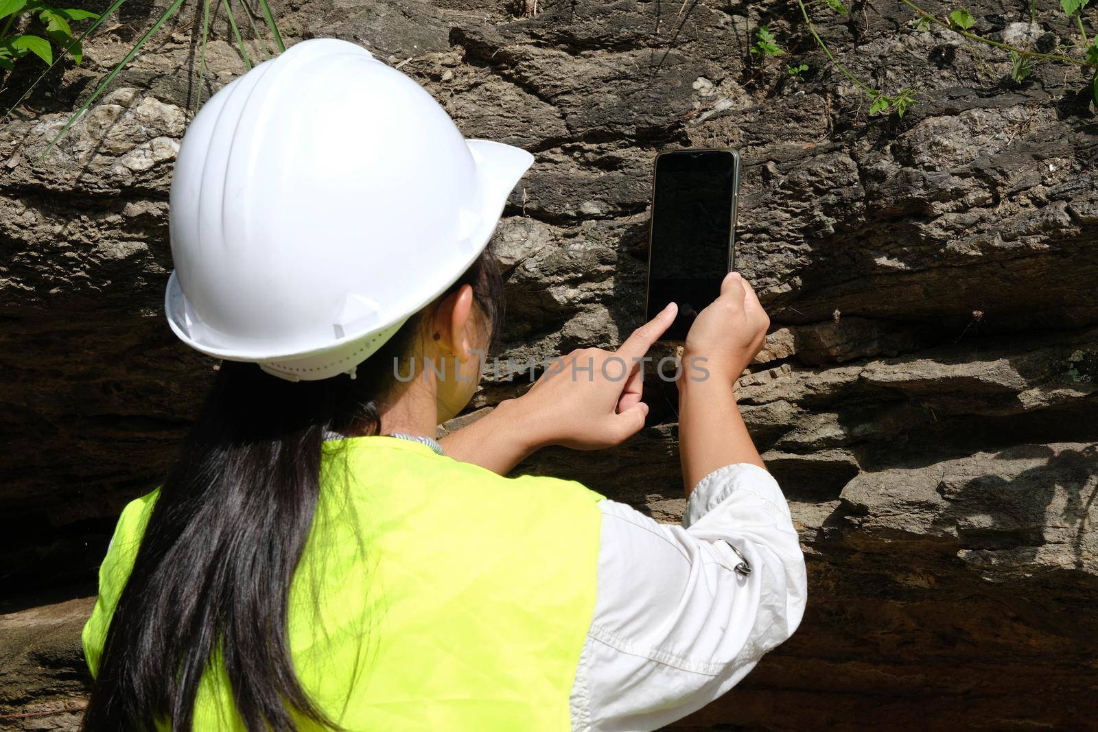 Asian female geologist researcher is analyzing rocks with a smartphone in a natural park. Exploration Geologist in the Field. Stone and ecology concept. by TEERASAK