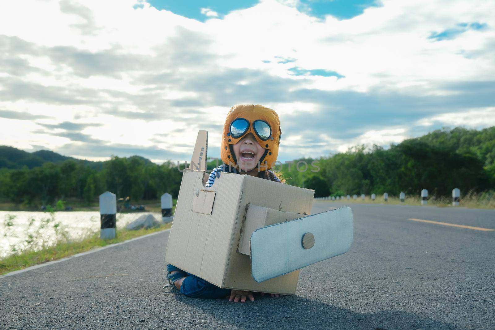 Cute dreamer little girl playing with cardboard planes on a lake road on a sunny day. Happy kid playing with cardboard plane against blue summer sky background. Childhood dream imagination concept.