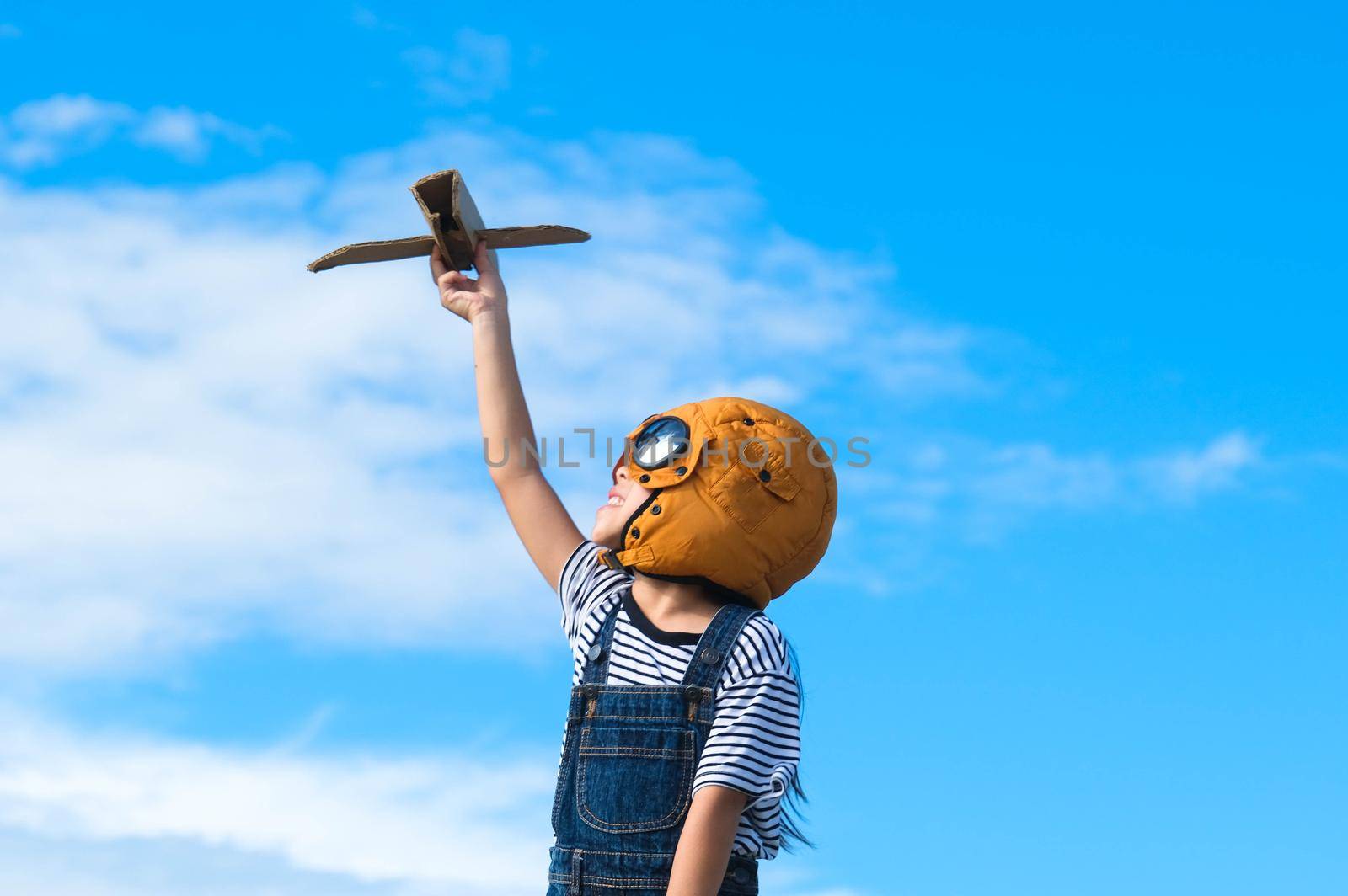 Cute little girl running through the meadow on a sunny day with a toy plane in hand. Happy kid playing with cardboard plane against blue summer sky background. Childhood dream imagination concept. by TEERASAK