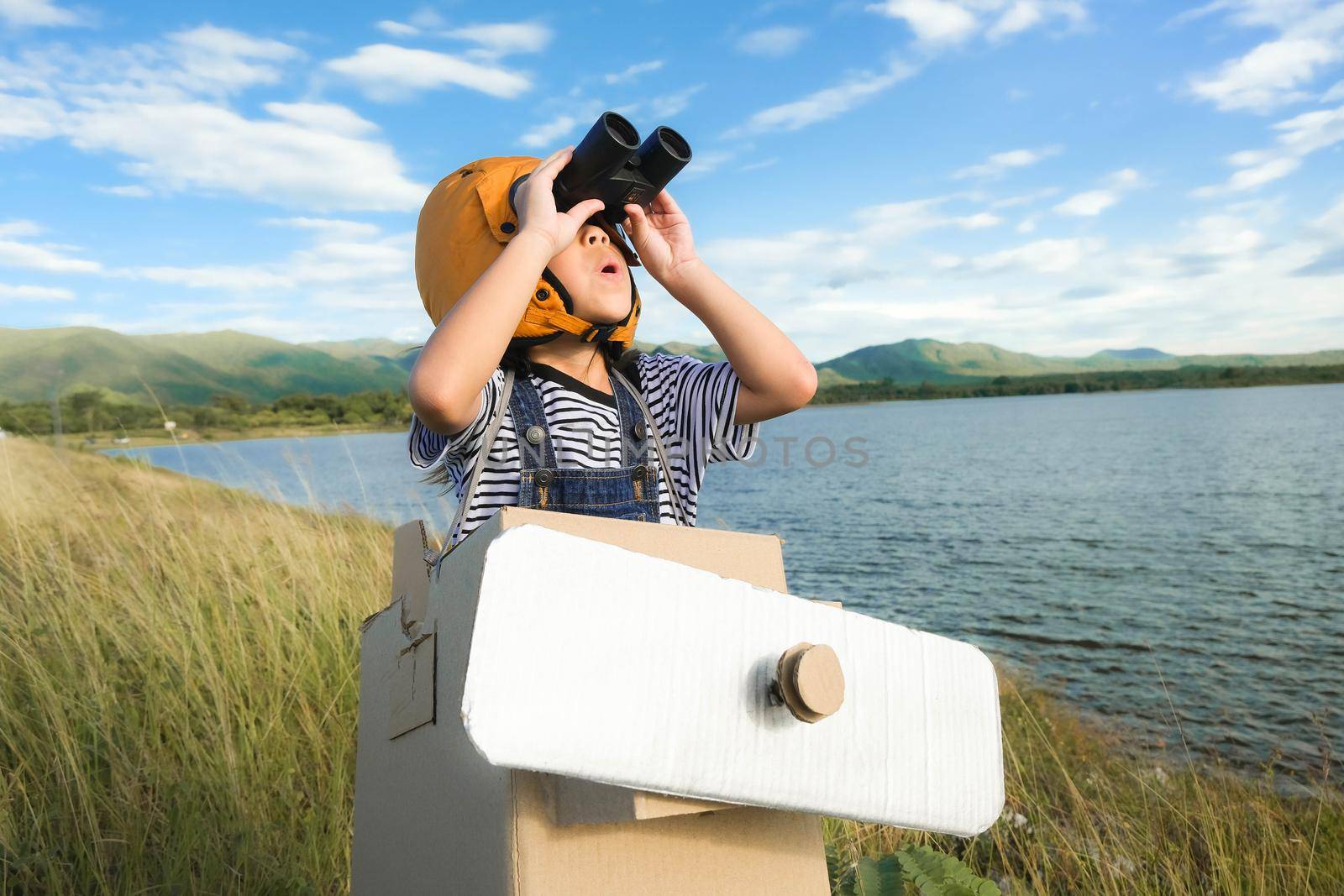 Cute dreamer little girl playing with cardboard planes and binoculars in a lakeside meadow on a sunny day. Childhood dream imagination concept.