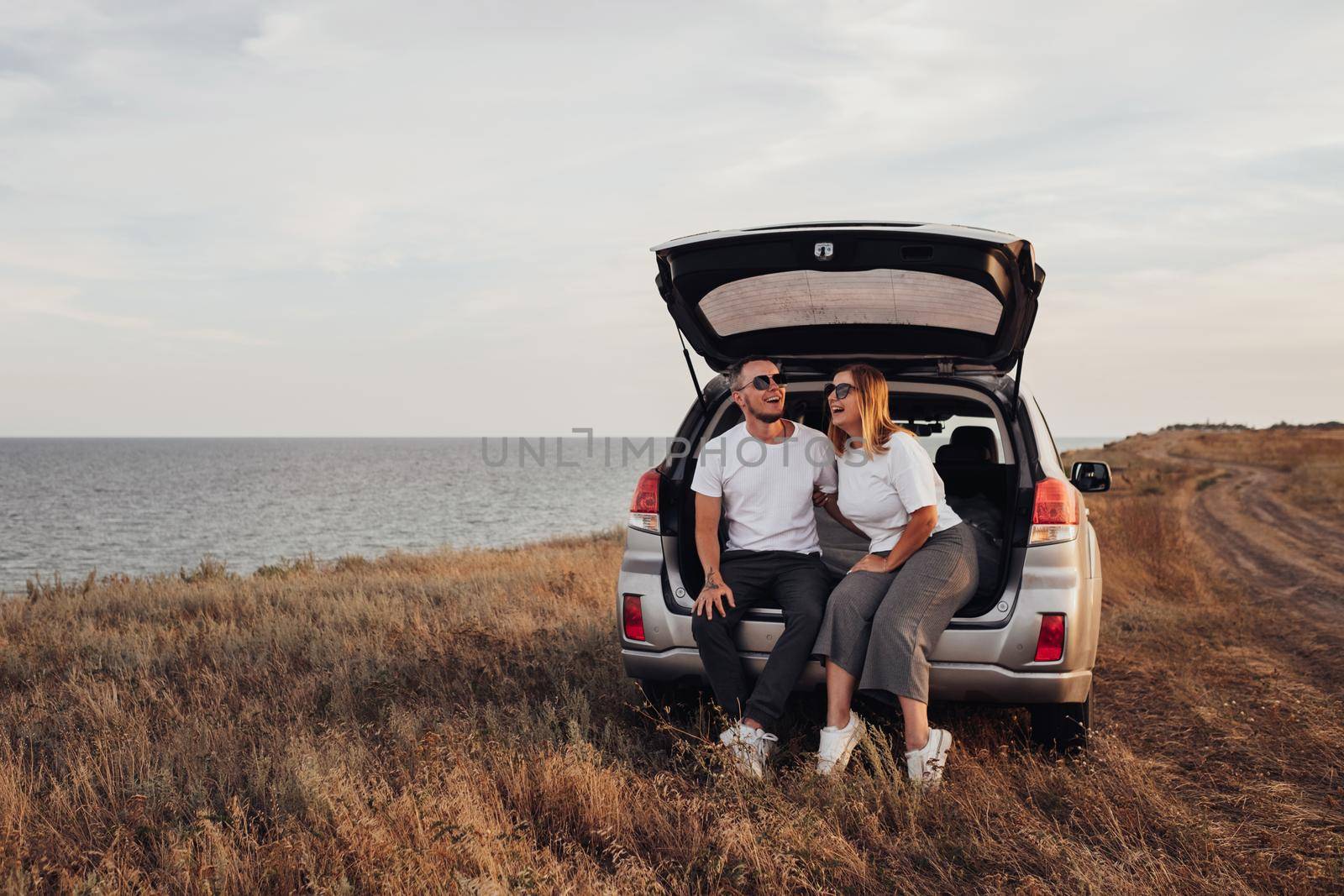 Happy Young Couple Sitting in Open Trunk of SUV Car, Man and Woman Enjoying Their Road Trip Along Seaside
