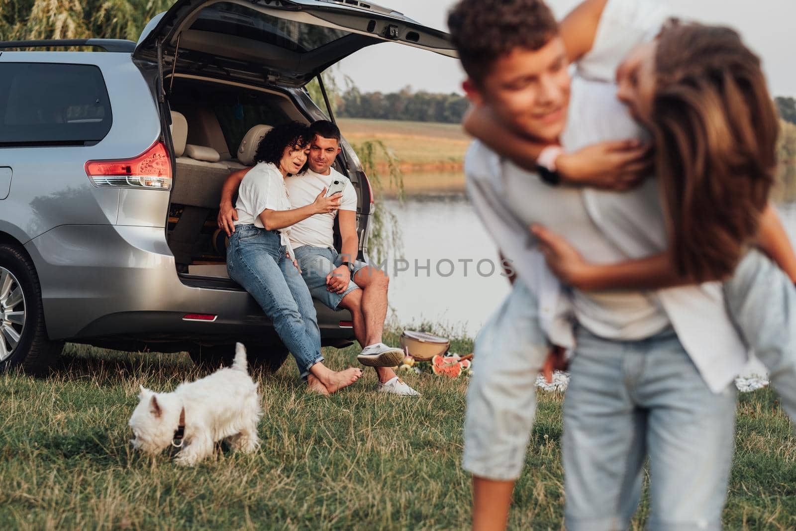 Happy Mother and Father Sitting in Trunk of Minivan Car While Children Playing with by Lake, Four Members Family and Pet Dog Having Weekend Picnic Outdoors