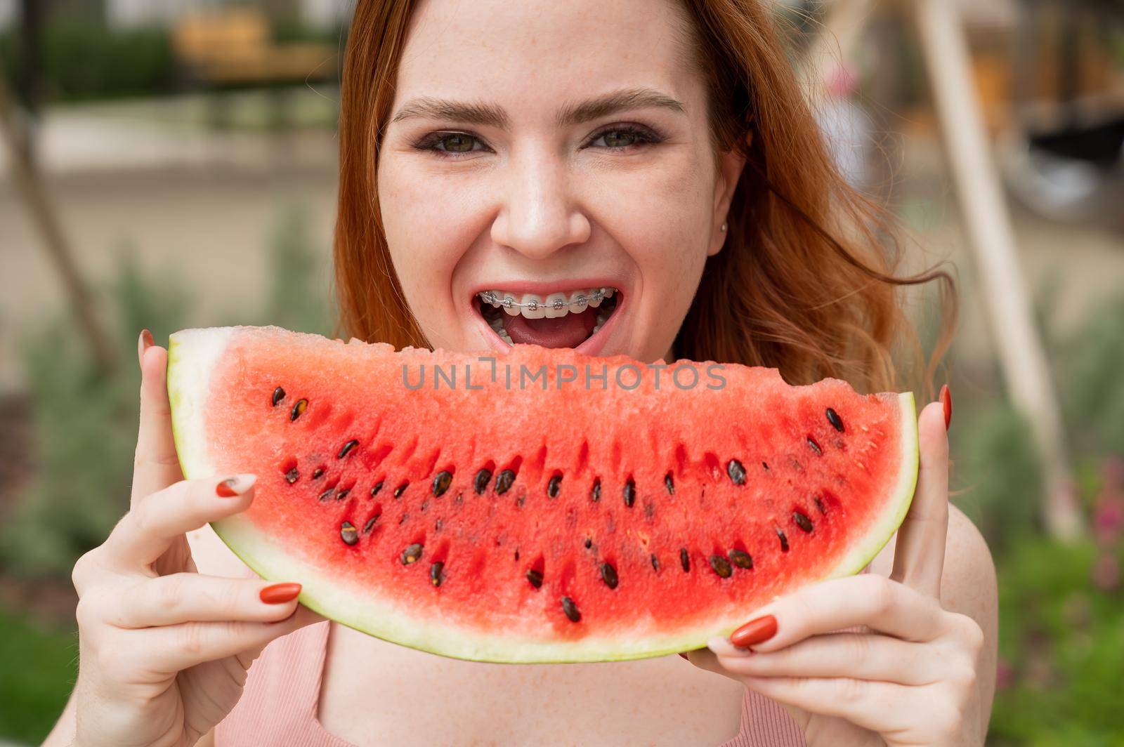 Beautiful red-haired woman smiling with braces and about to eat a slice of watermelon outdoors in summer.